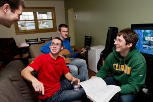 Four male Clarkson students hang out together in a common lounge of a residence hall, including one who plays the guitar. 