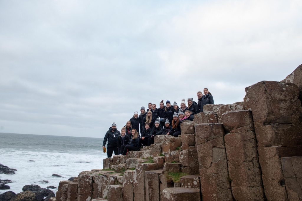 A group photo with them standing and sitting on the edge of a cliff with the ocean in the background