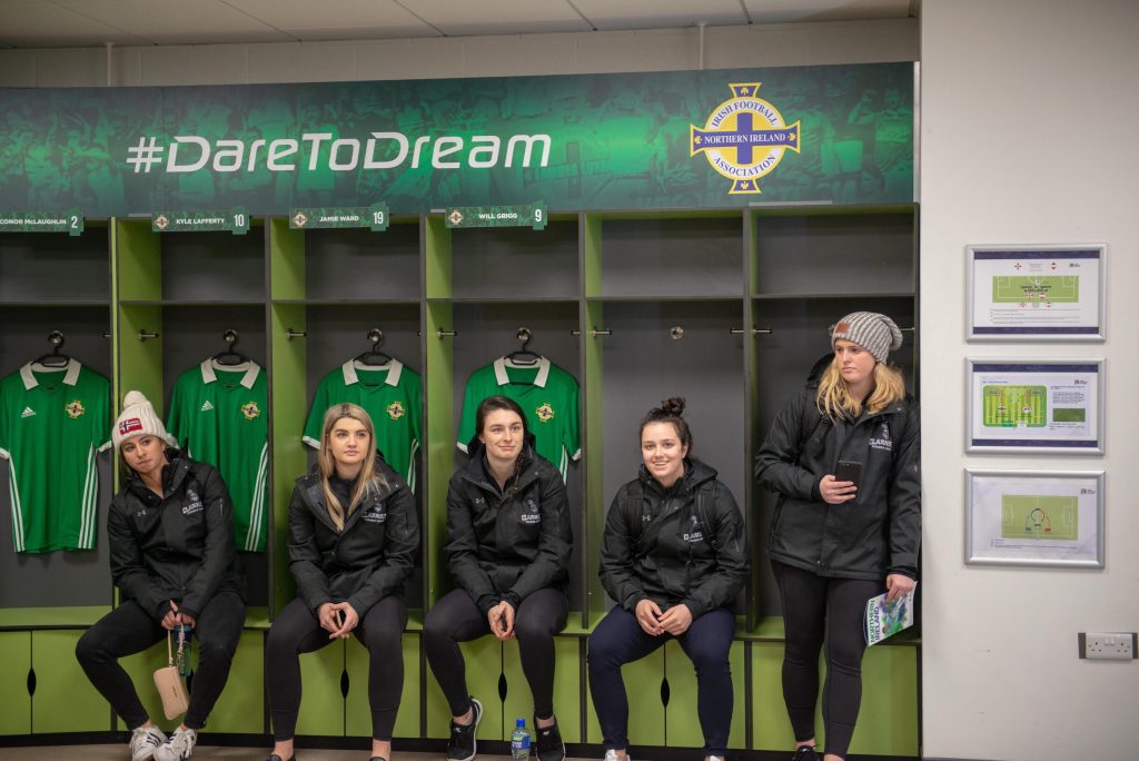 A group of women sitting on a green sports bench with the sign above reading Dare to dream