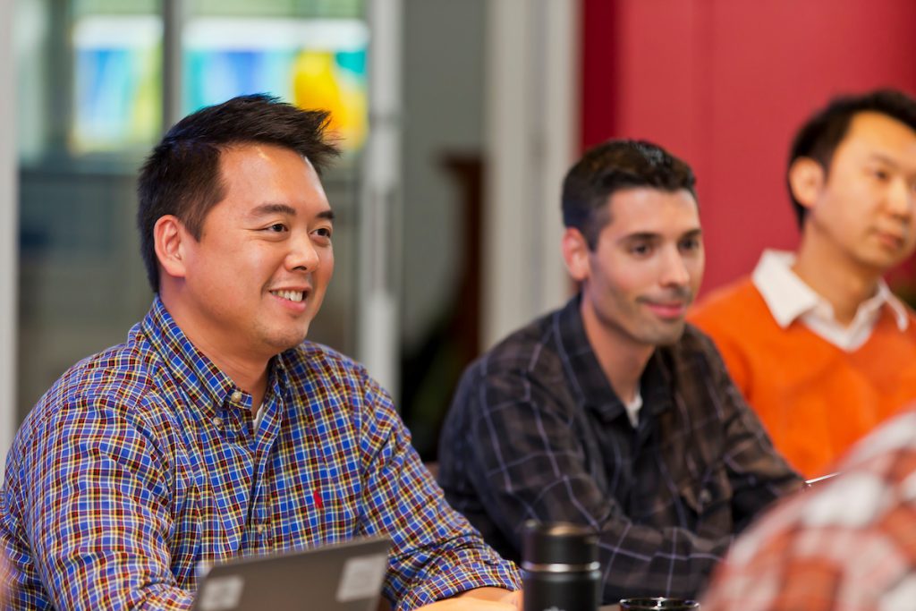 Three men sitting at a table listening to a lecture