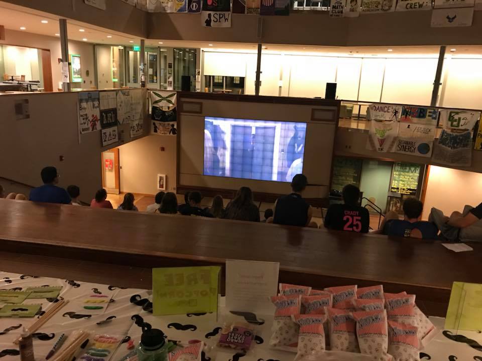 A dimly light student center with students sitting to watch the projector screen that is playing something