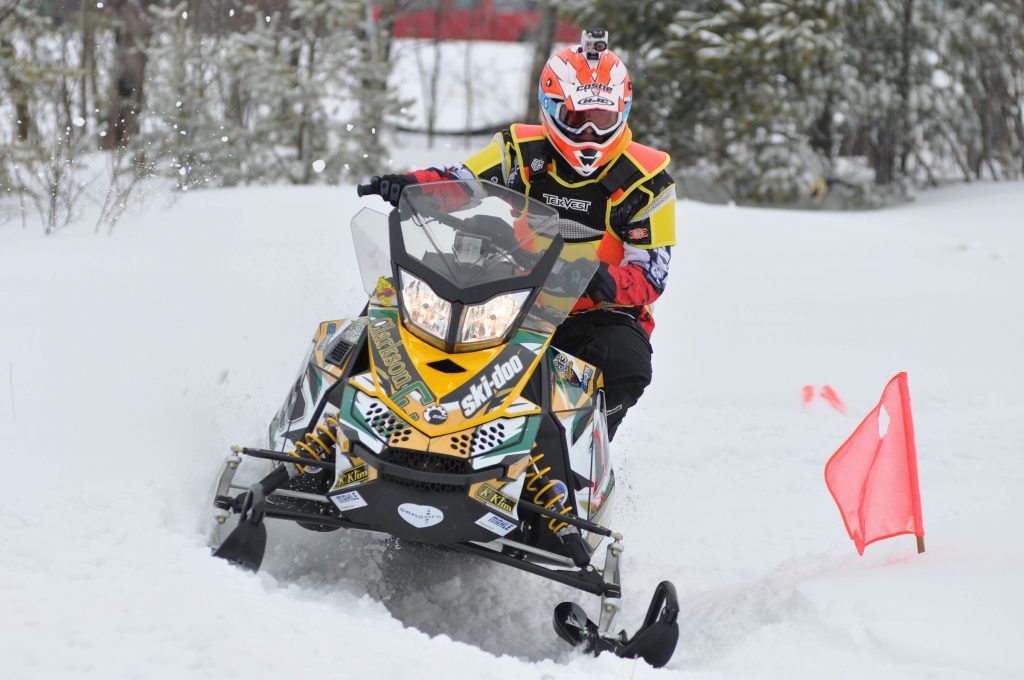 A person in a helmet and snow suit on a yellow snowmobile with orange flags marking the trail