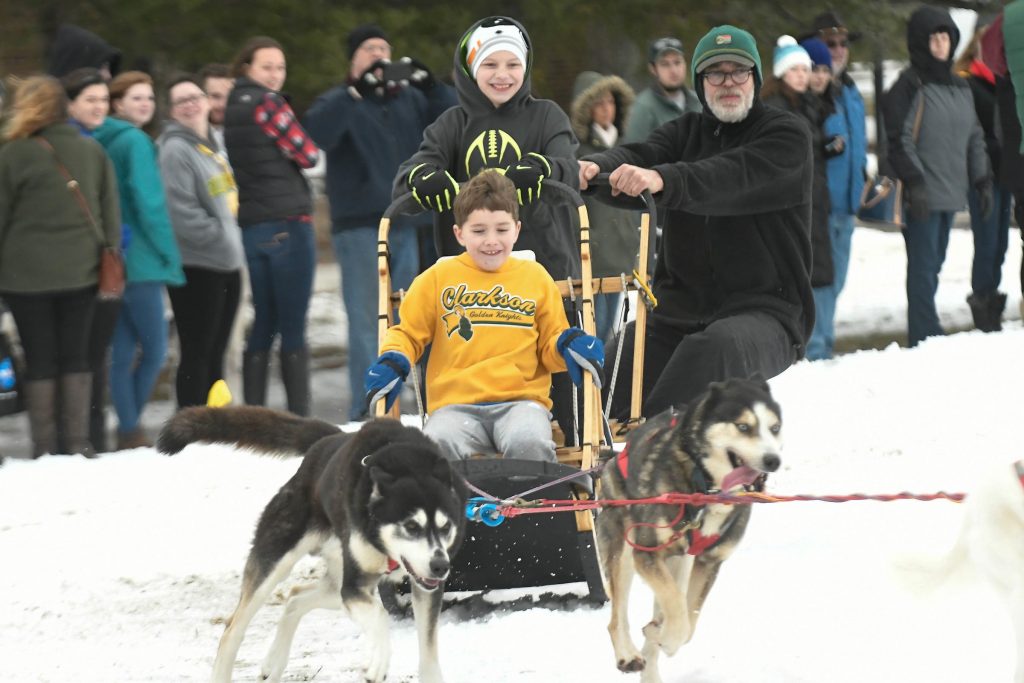 A child in the sit of a dog sled with another person holding onto the back and a man steering. Two dogs are running at full speed