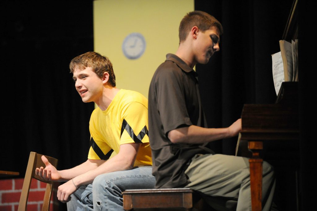 Two students sitting on a piano bench one playing and the other facing the other direction 
