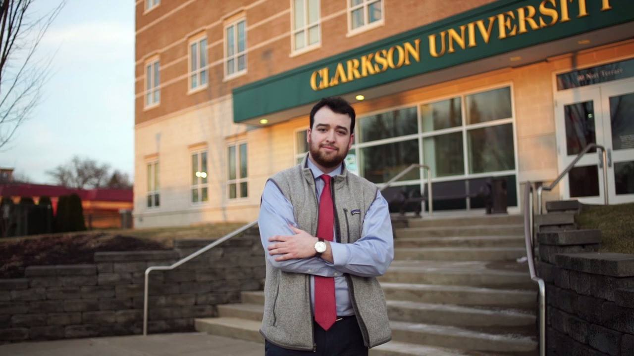 A man standing in front of the Clarkson University Capital Region Campus with a sign that reads Clarkson University