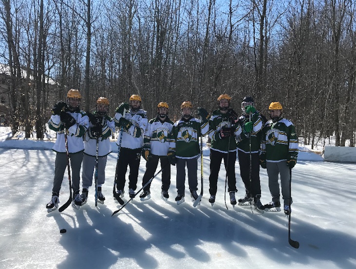 Several members of Clarkson University Men's Club Hockey stands on an outdoor hockey rink wearing helmets and holding hockey sticks. 