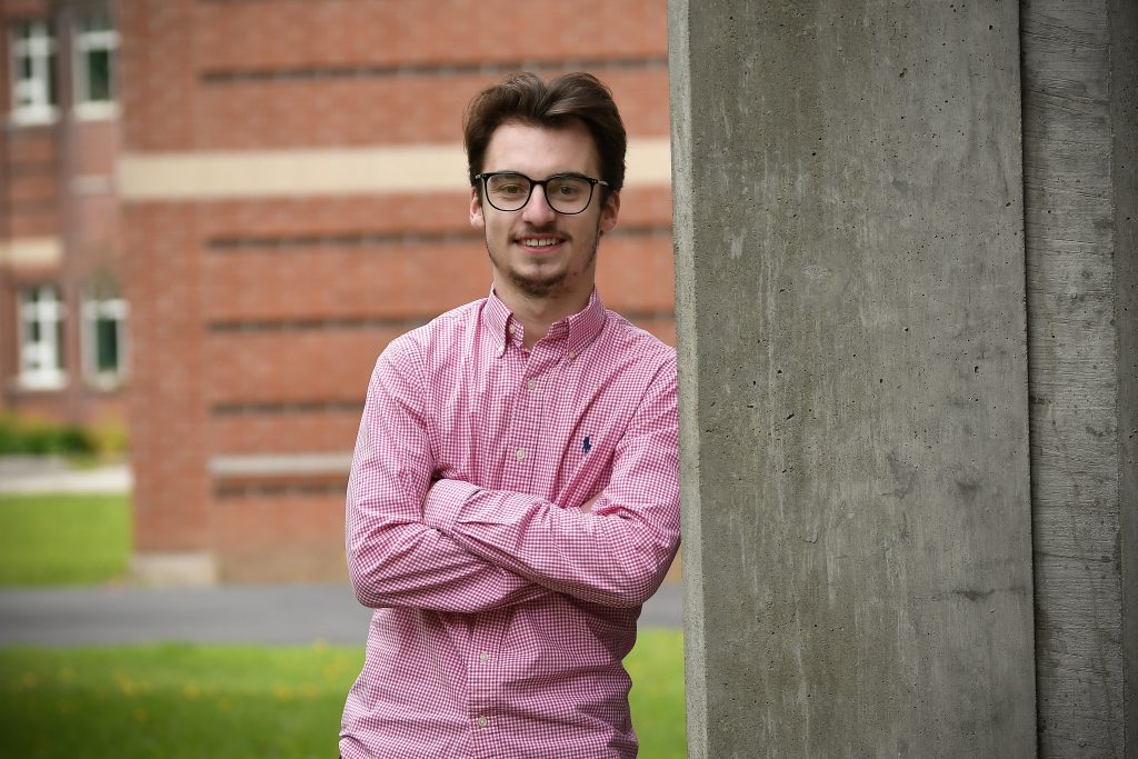 Nick Kozsan, wearing a pink shirt and glasses, poses with his arms crossed on Clarkson's campus. 