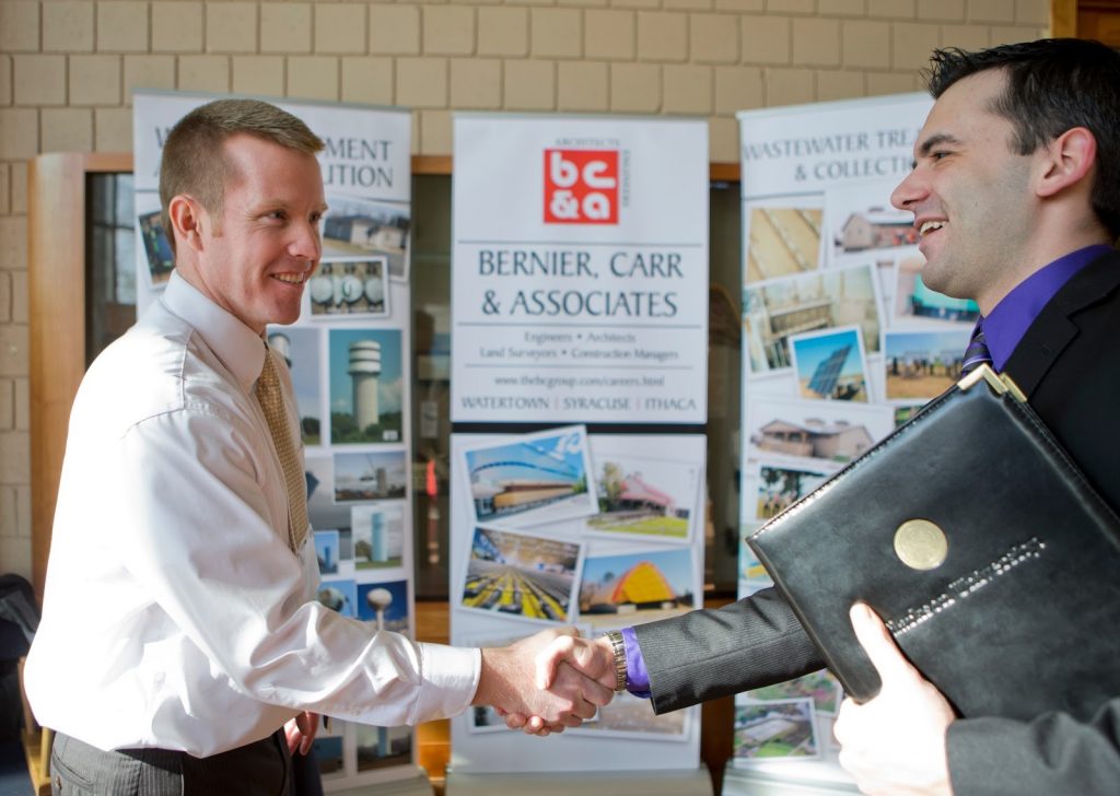 A student holding a resume folder shaking hands with a man standing in front of a display booth