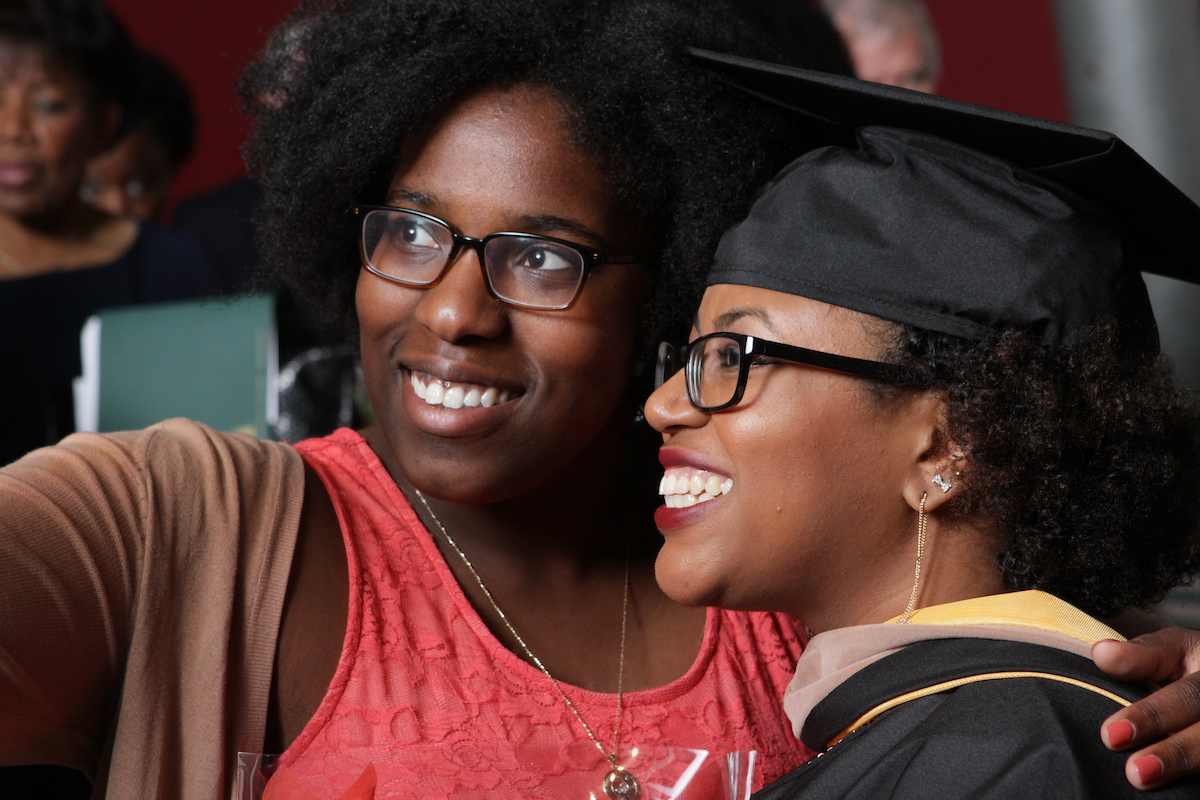 A student wearing regalia poses for a selfie with a friend.