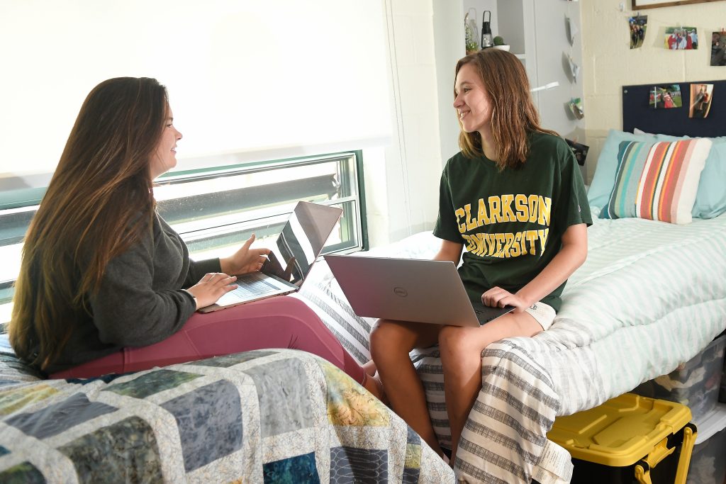 Two Clarkson students relax in their beds in their Cubley House residence hall room while holding laptops.