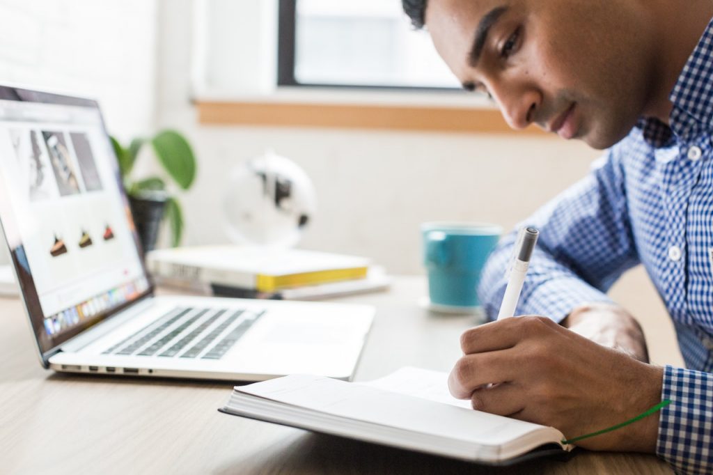 A man writes in a notebook with his laptop in the background.