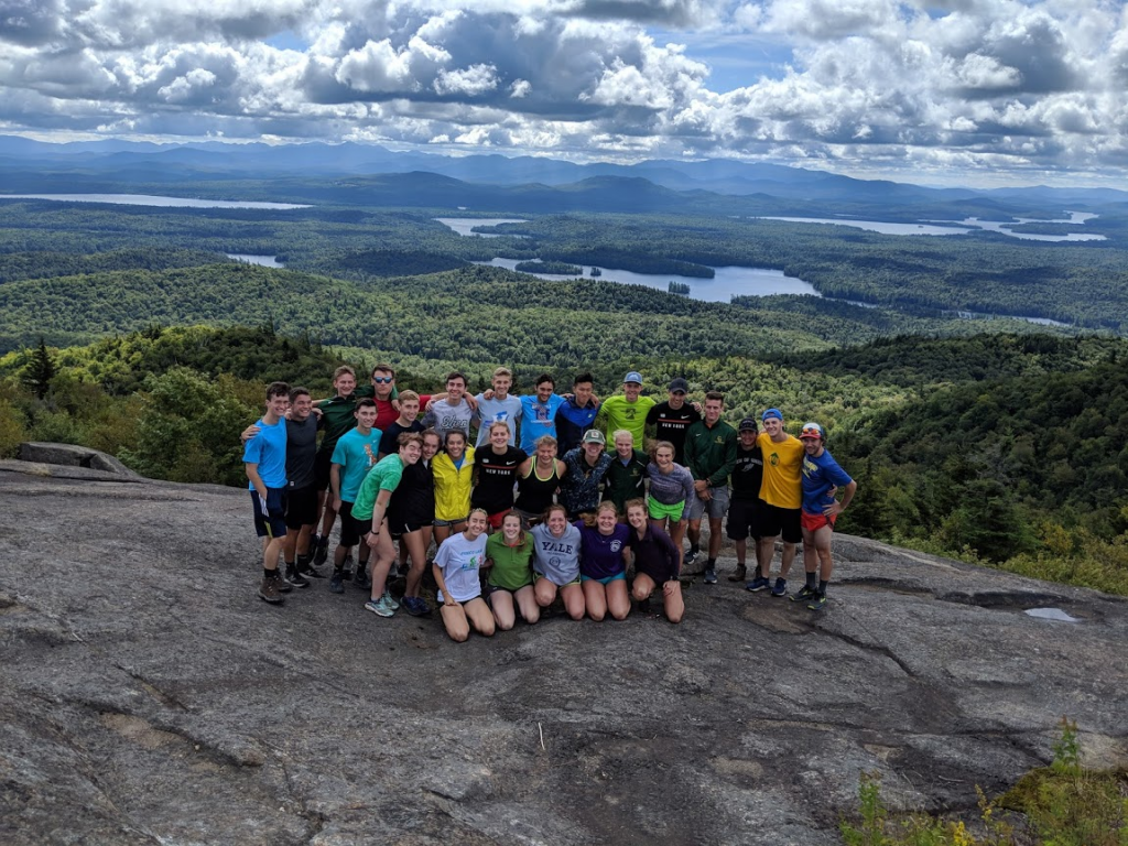 Group of Clarkson students standing together on the peak of a mountain overlooking woods and lakes