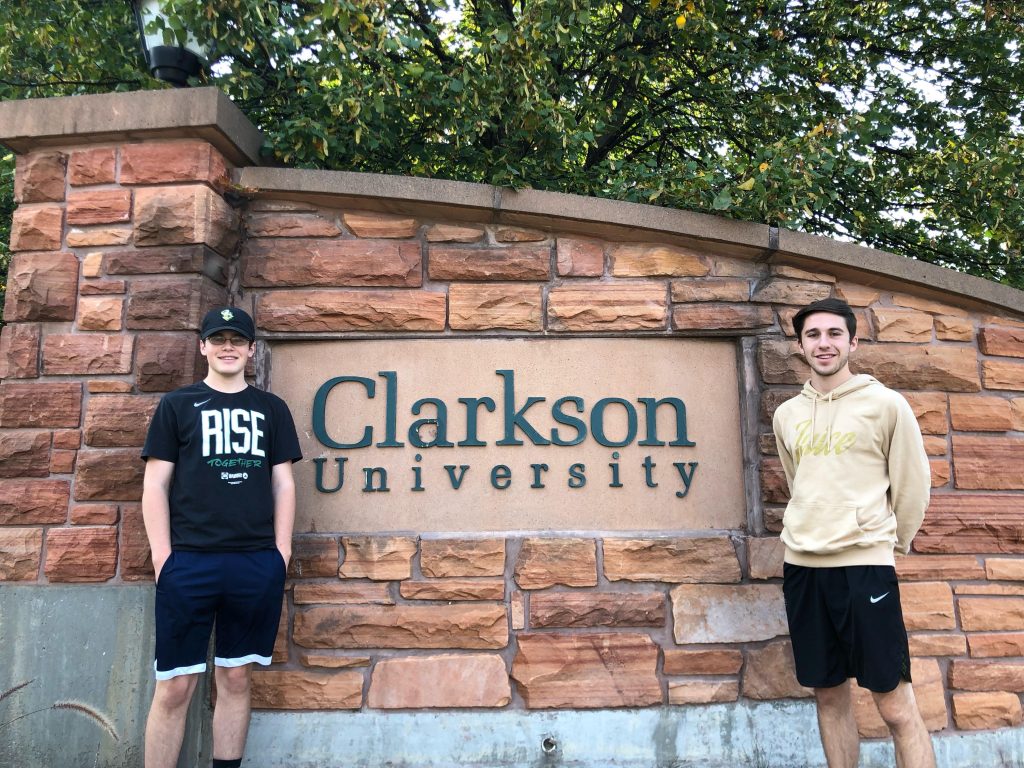 Two students pose on either side of a large Clarkson University sign on a piece of stone that makes up the welcoming entrance to campus. 