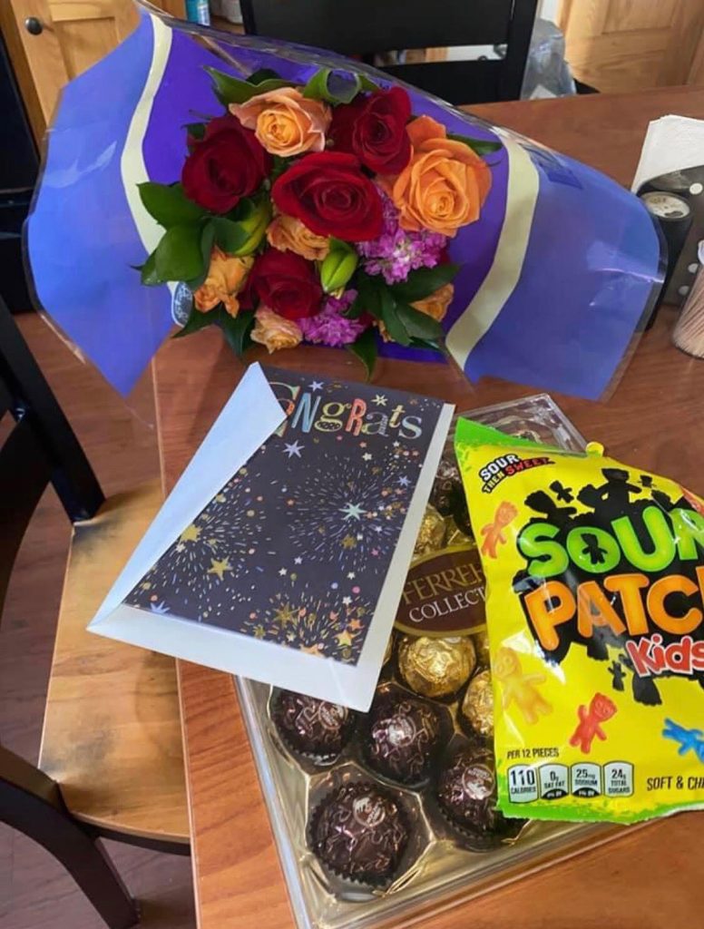 flowers, a congratulatory card, chocolates and some sour candies lay on top of a desk. 