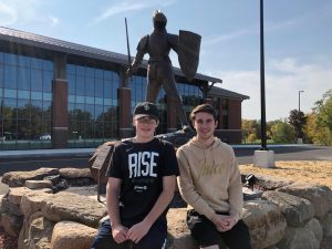 Two students sit on a large stone posing in front of a statue of a knight.