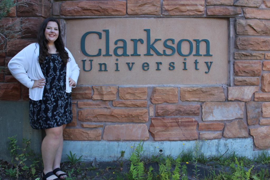 A person standing next to the stone sign that reads Clarkson University 