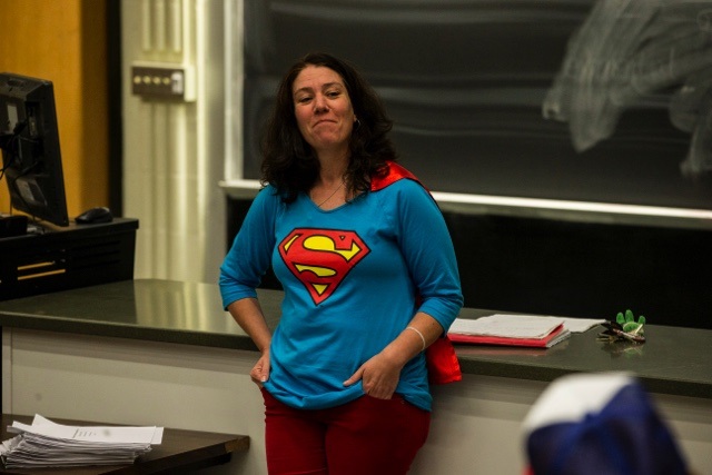 a university professor with long dark hair interacts with her class wearing a blue shirt with a Superman logo with a short red cape. 