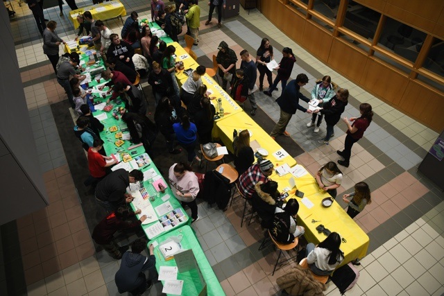 A photo taken from above depicts two long tables, one yellow and one green, littered with papers and activity props as dozens of students gather around each table. 