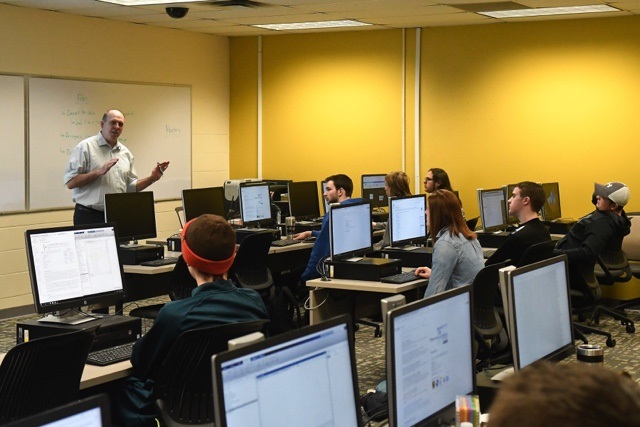 A university professor animates his words with his hands as he addresses a classroom full of students, who are all sitting behind their own desktop computer. 
