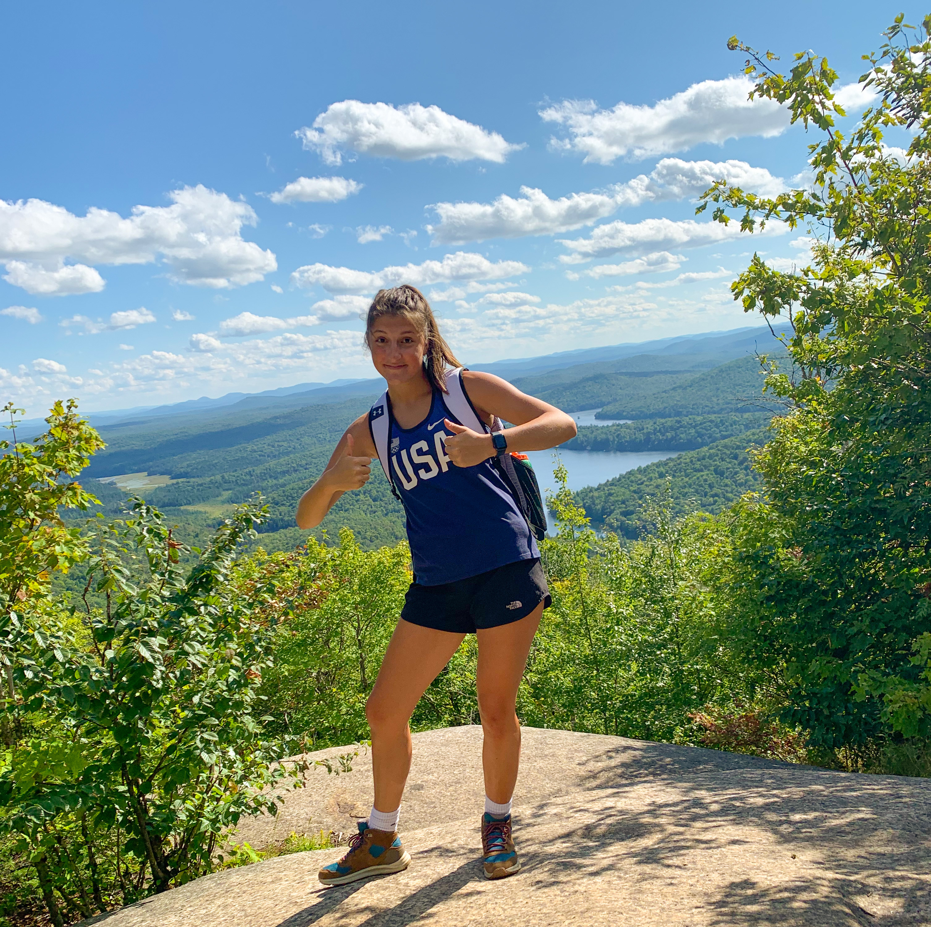 A student in a blue tanktop poses with both thumbs up on a mountaintop on a sunny day.