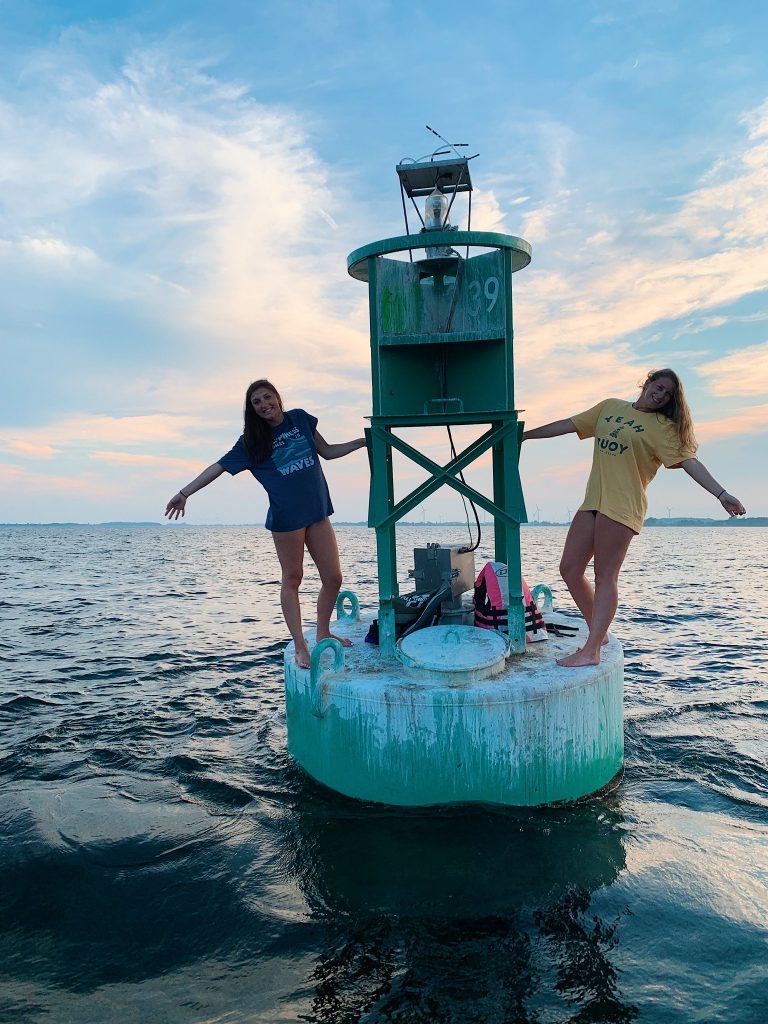 Two students stand on either side of a large buoy on a river. 