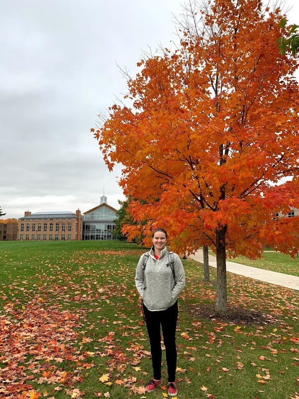 A woman in black pants and a gray pullover poses in front of a tree with red leaves. A large brick and glass building can be seen far in the background. 