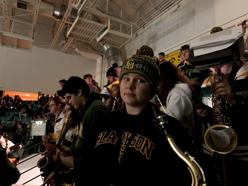 A student stands posed with her saxophone in a winter hat surrounded by band members at a hockey game. 