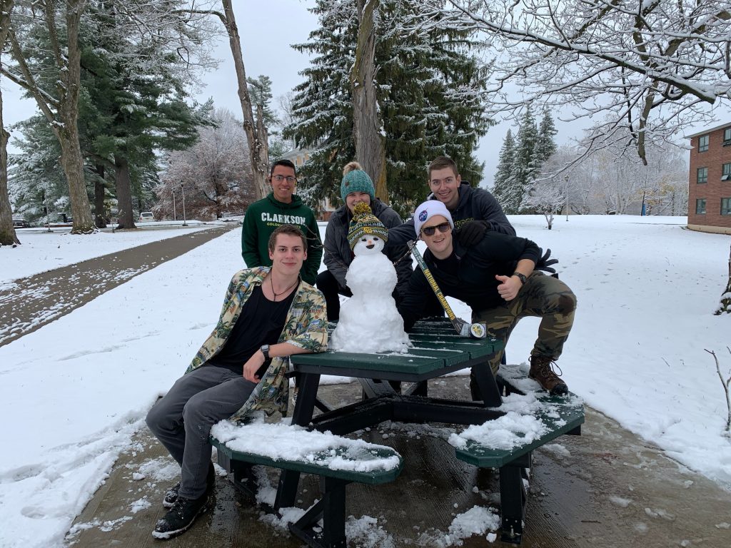 Five students pose around a small snowman situated on a picnic table. 