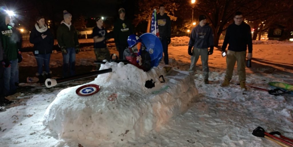Several students stand around a tank made of snow in an open space at night. 