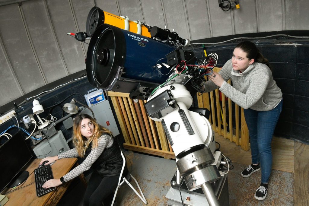 A student adjusts a large telescope she stands behind, while another student is seated at a computer next to the telescope. 