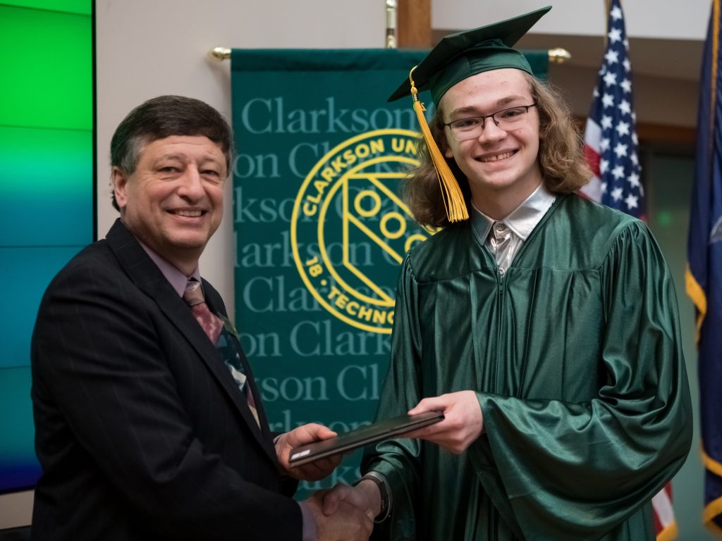 A man in a suite passing over a diploma holder to a person in a green cap and gown