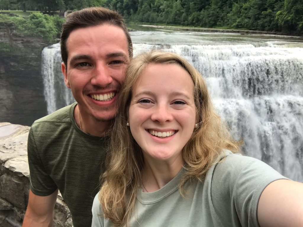 A couple smiling into the camera with a waterfall as the background 