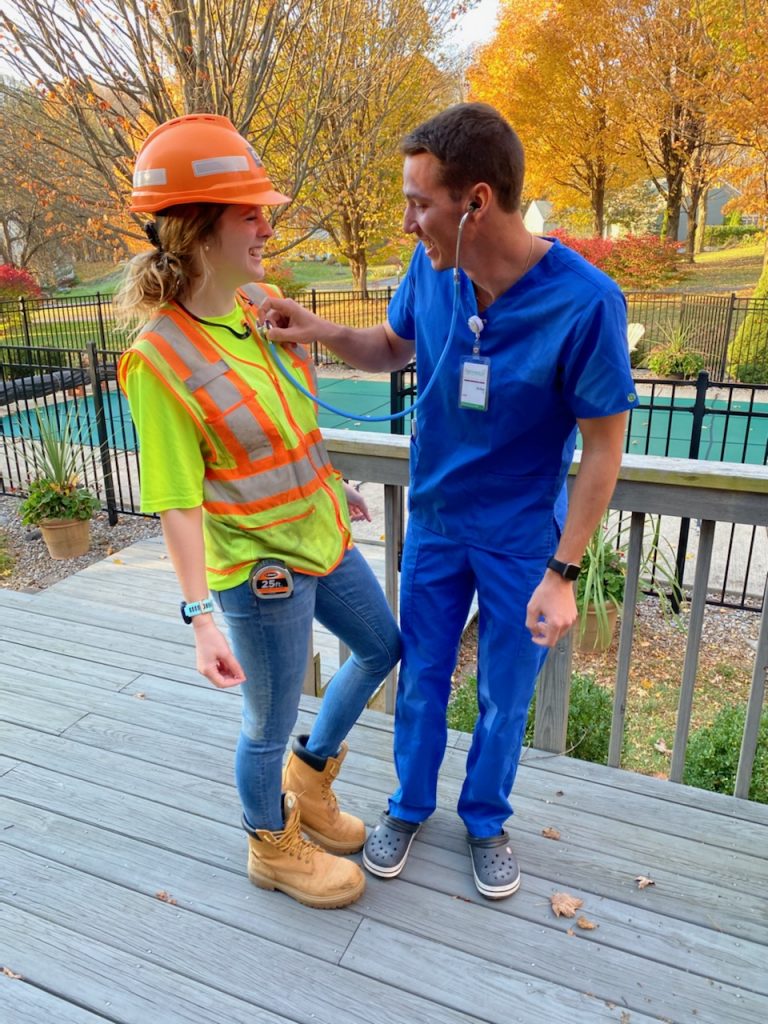 A man in scrubs holding a stethoscope up to a women in a construction outfit