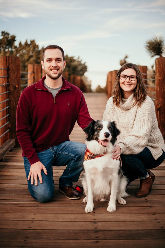 A couple squatting on a bridge with a dog in between in a plaid bandanna 