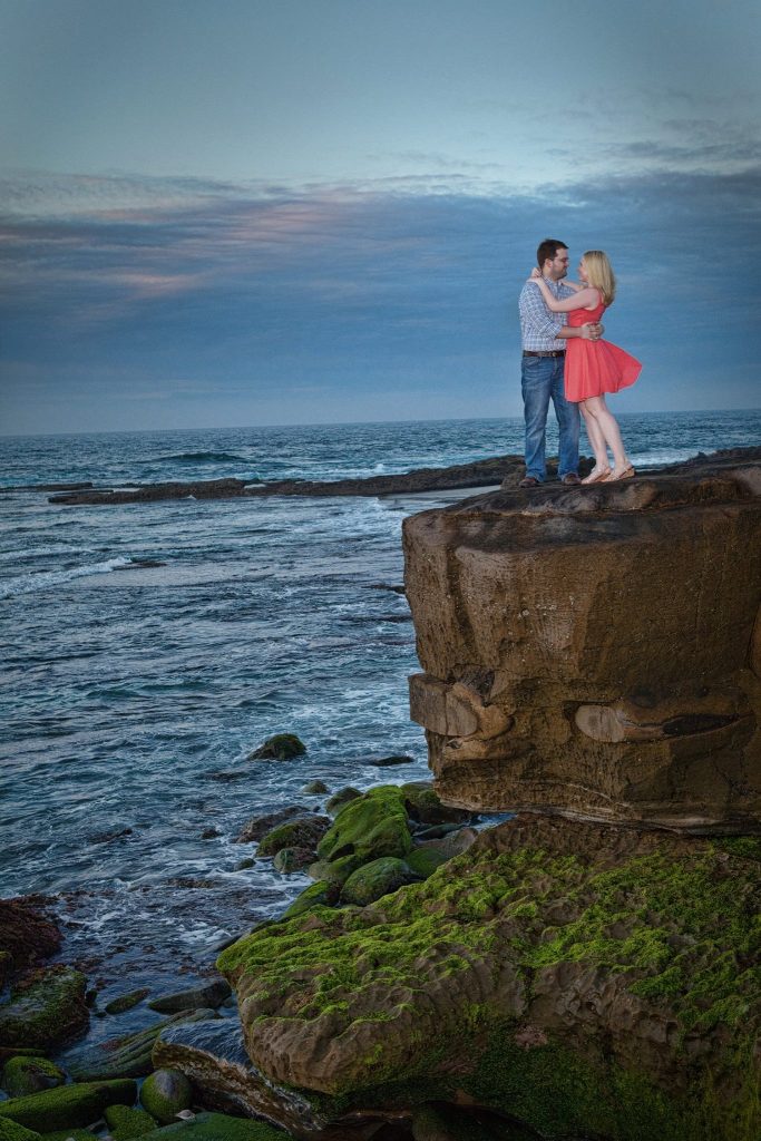 A man and woman pose while looking at each other on a large rock overlooking the ocean. 