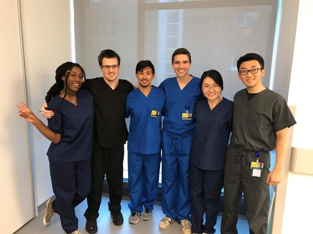 Six medical students smile at the camera as they pose for a photo in their scrubs. 