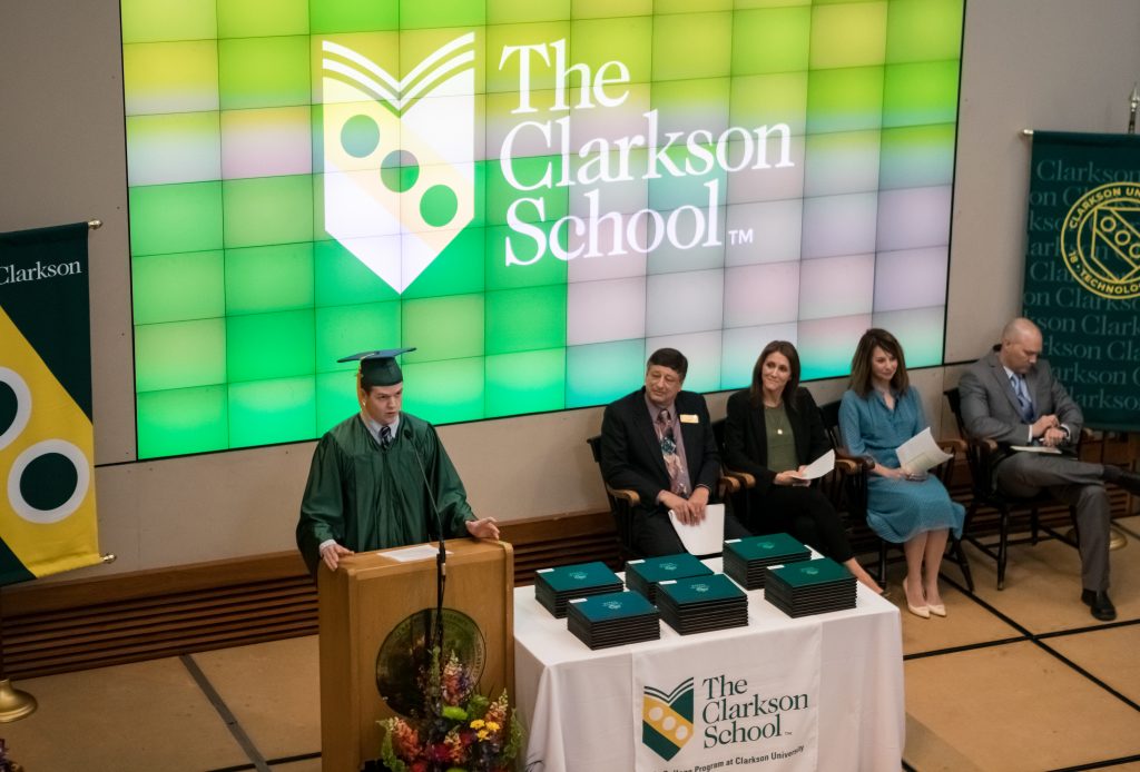 A Clarkson School student standing in graduation regalia behind a podium, during the TCS Commencement