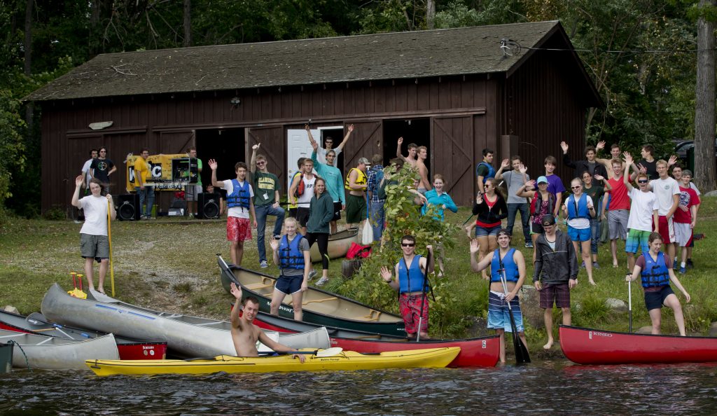 Looking from the water at a large group of students  congregating on shore with some in canoes, during Clarkson University Outing Club's Canoefest