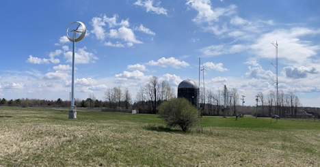 a ducted wind turbine stands on the left of the photo in a mostly open field with a few scattered trees and a silo in the background. 