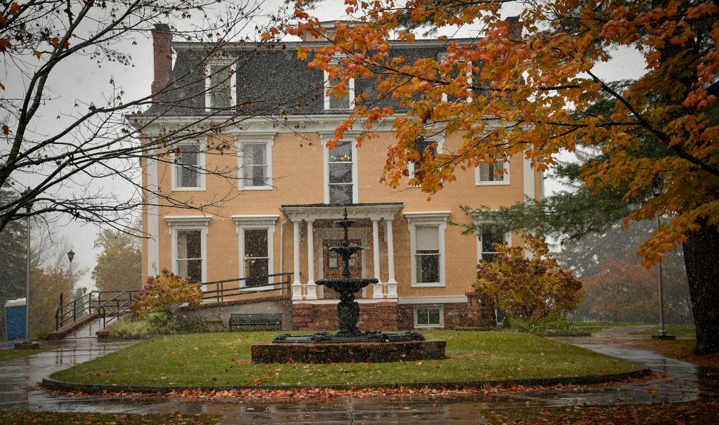 A large yellow building is centered behind a water fountain as trees with changing leaves hang in front on a rainy day. 