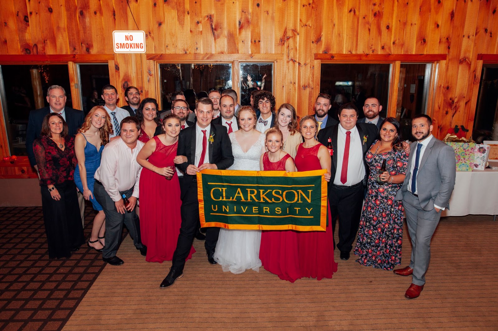The Wedding party and guests standing behind a bride and groom while holding up a Clarkson University flag