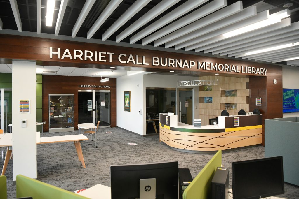 Large white lettering spells out "Harriet Call Burnap Memorial Library" across a brown entryway, which overhands a circulation desk surrounded by other desks and chairs. 