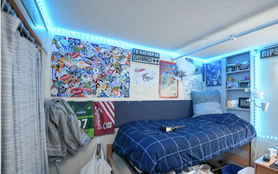 A student's dorm room is complete with a raised bed covered in a blue comforter, with several skiing posters on the wall which are surrounded by a string of blue LED lights. 