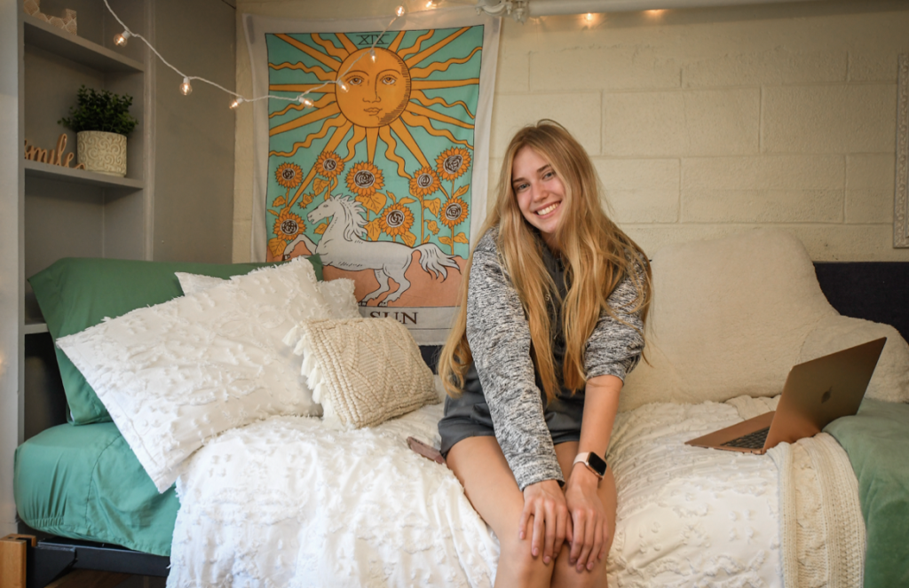 A student sits smiling on her green and white bedding in her dorm room, with a poster in the background featuring a sun, sunflowers, and a horse. Her laptop is open on the bed beside her. 