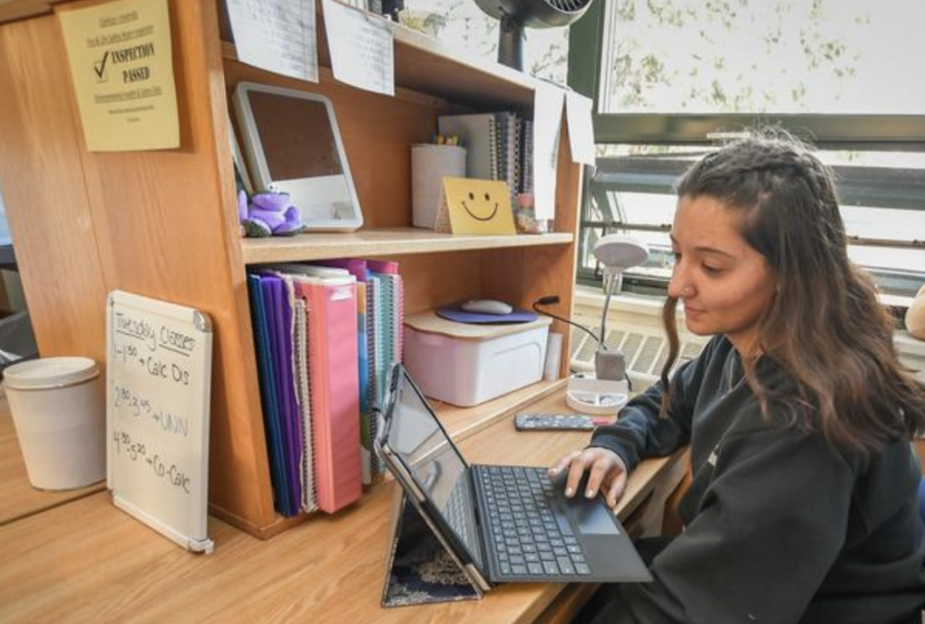 A student sits at her desk in her dorm room scrolling on her laptop.