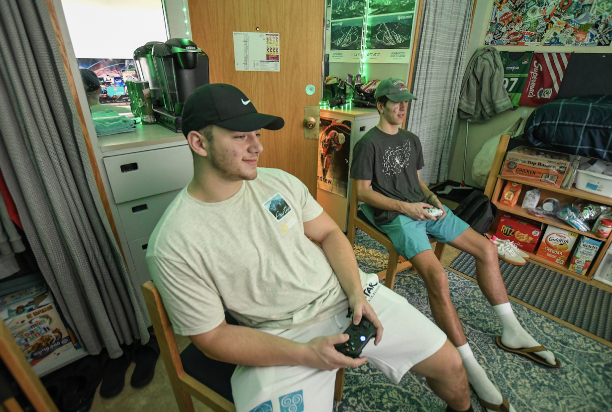 Two students hold video game controllers while seated on wooden chairs in their dorm room.
