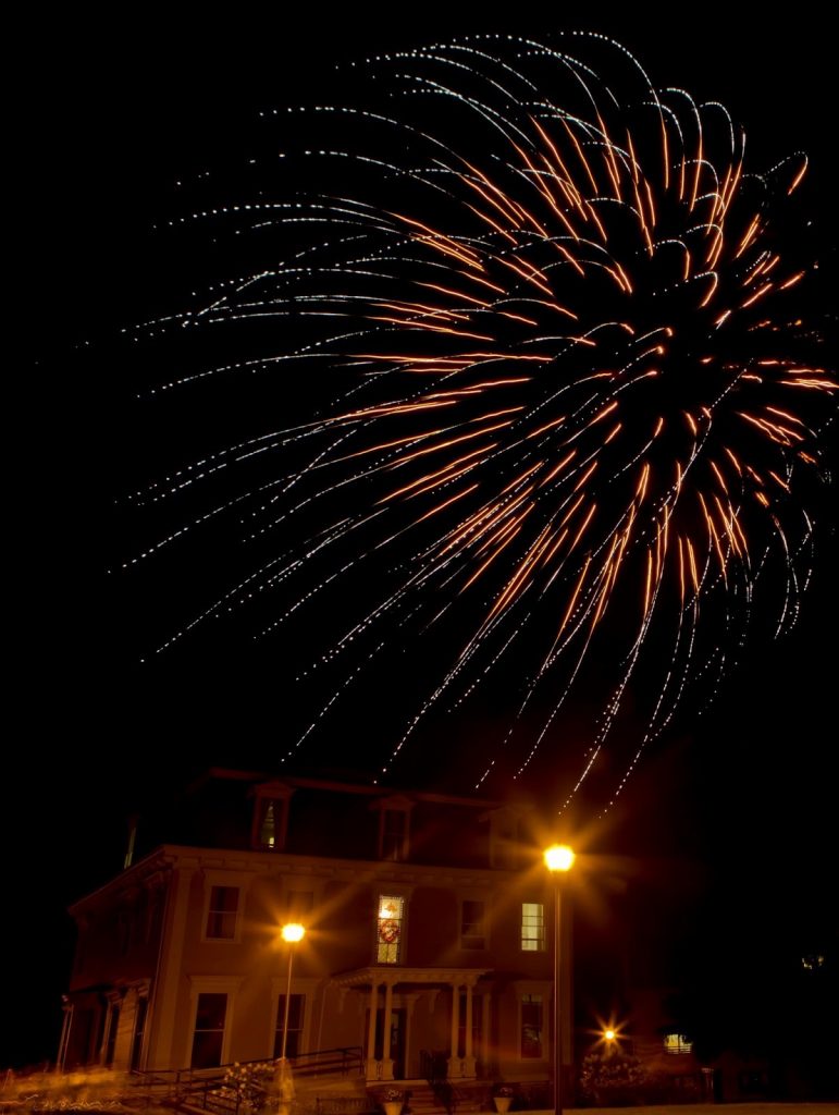 Fireworks of yellow and white shine over the Holcroft house with yellow street lights on 