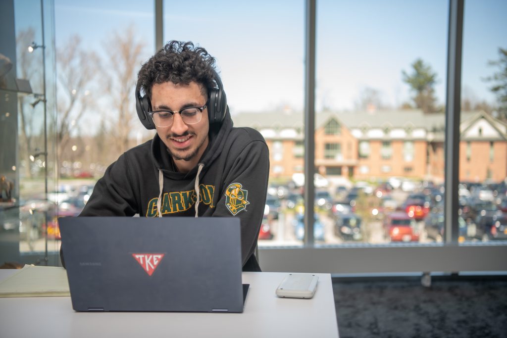 a student in glasses and a Clarkson hoodie uses his headphones and laptop in a common space on campus. 