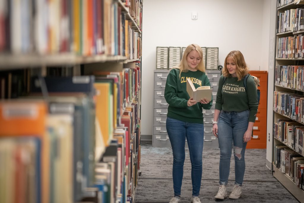 Two young women peruse a book in an aisle of the library, surrounded by books. 