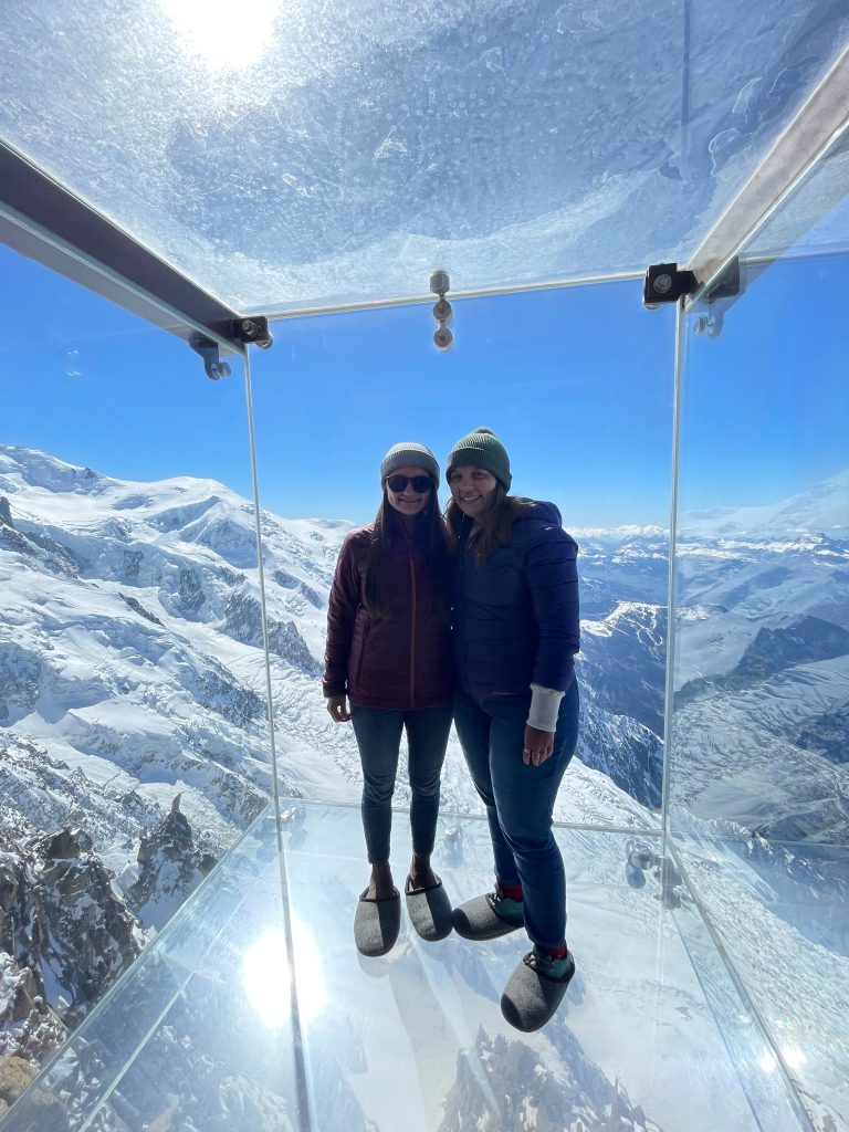 Two people standing in a clear glass box over mountains 